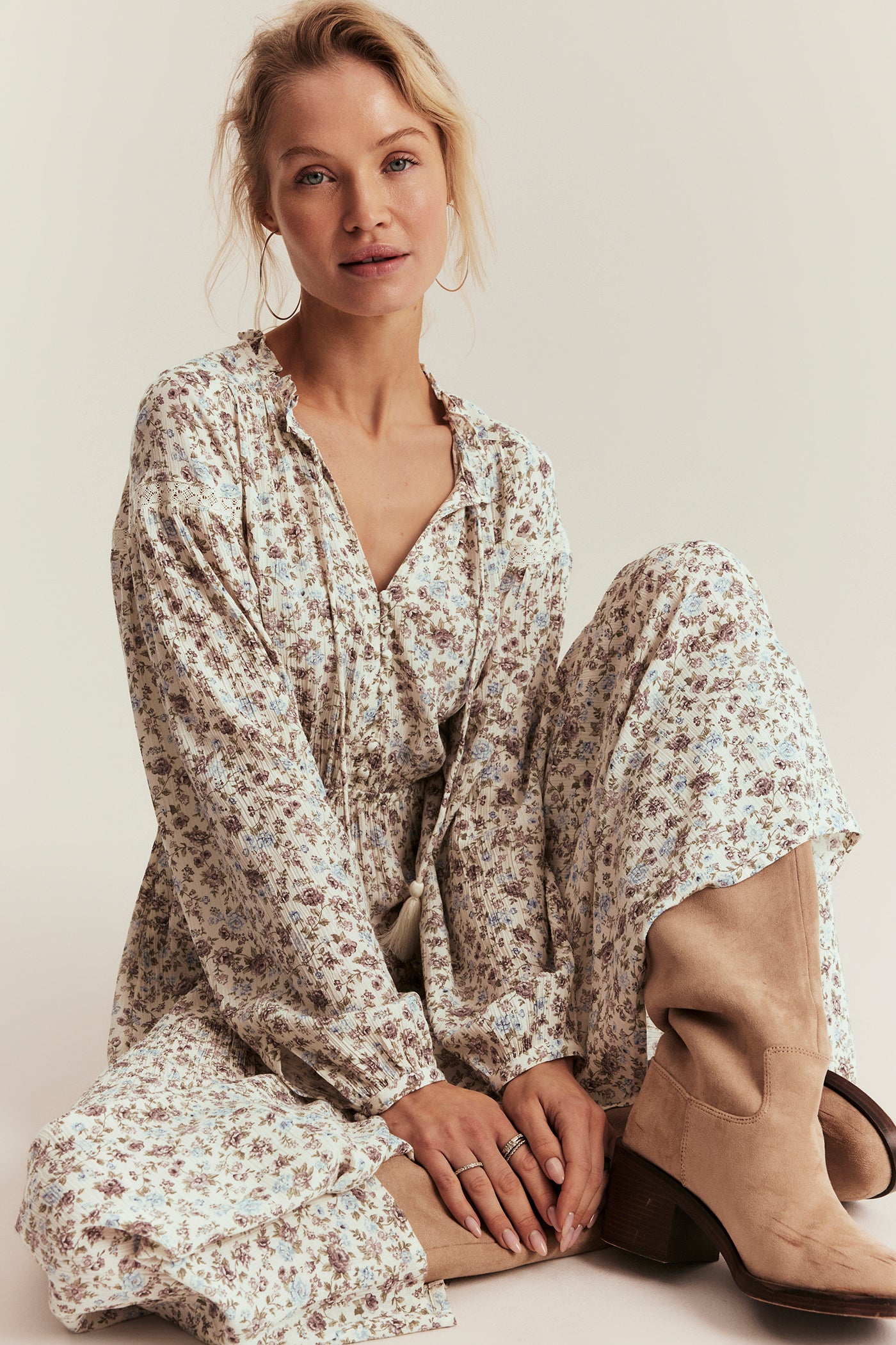 Woman wearing a floral dress and beige boots sitting on the floor against a plain background